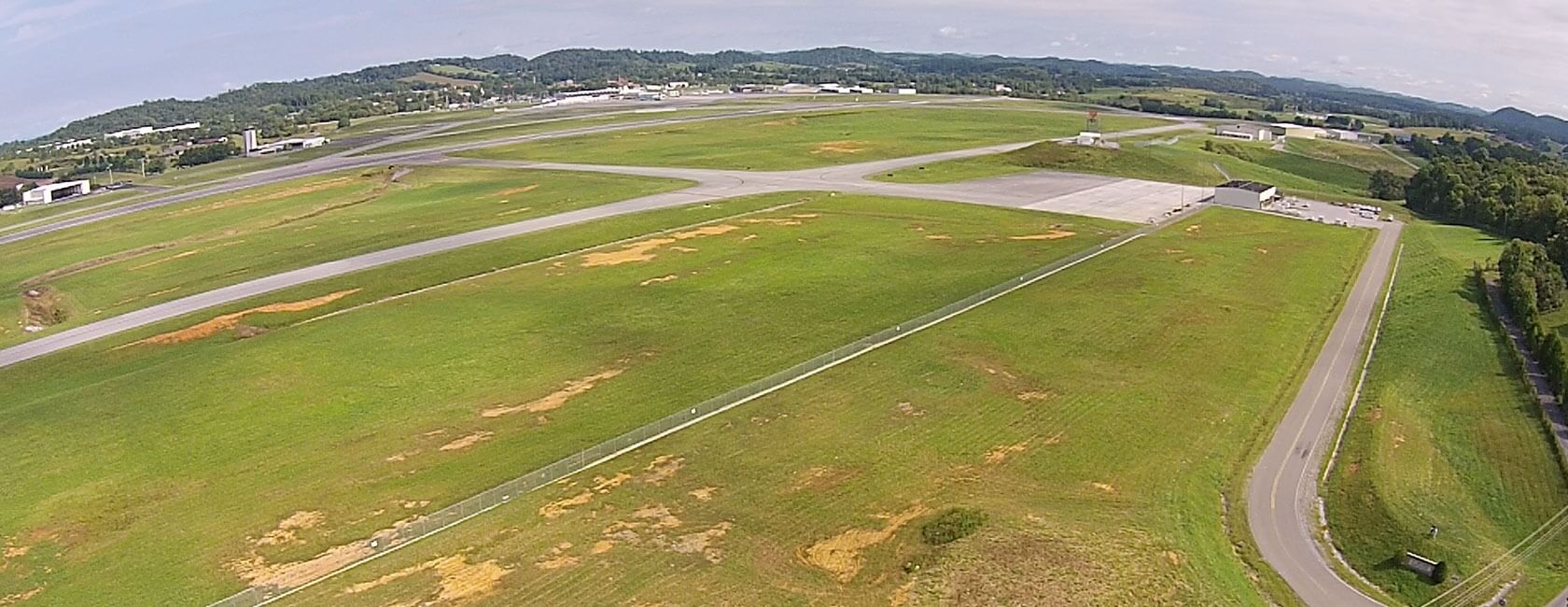 Aerial view of a small airport with intersecting runways, taxiways, hangars, and green grassy areas surrounded by trees and distant hills under a partly cloudy sky.