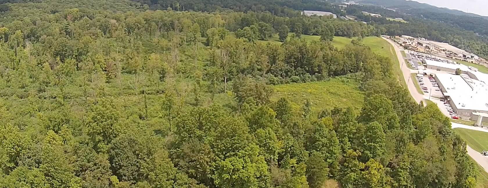 Aerial view of a mixed forest with dense green trees, open grassy areas, and industrial buildings with parking lots along a road to the right. Hilly landscape and more forest are visible in the background.