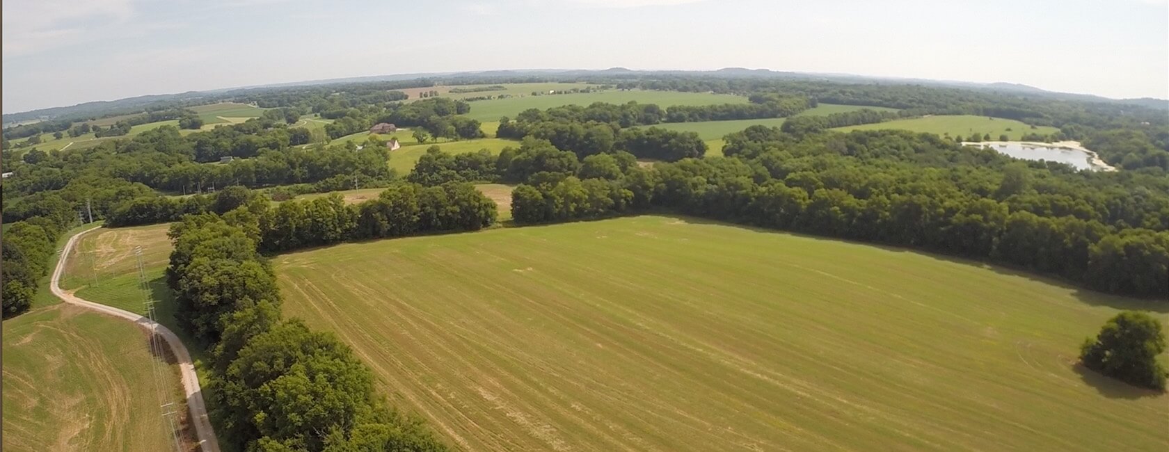 Aerial view of a large, open green field bordered by dense trees, with a winding road on the left and distant rolling hills under a clear sky.