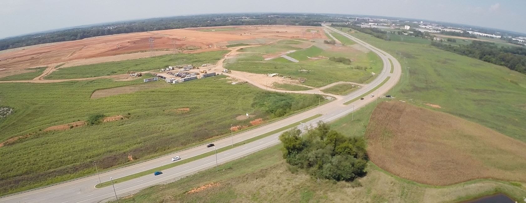 Aerial view of a rural highway curving through green fields, with a construction site and large cleared land area in the background under a partly cloudy sky. Several vehicles are visible on the road.