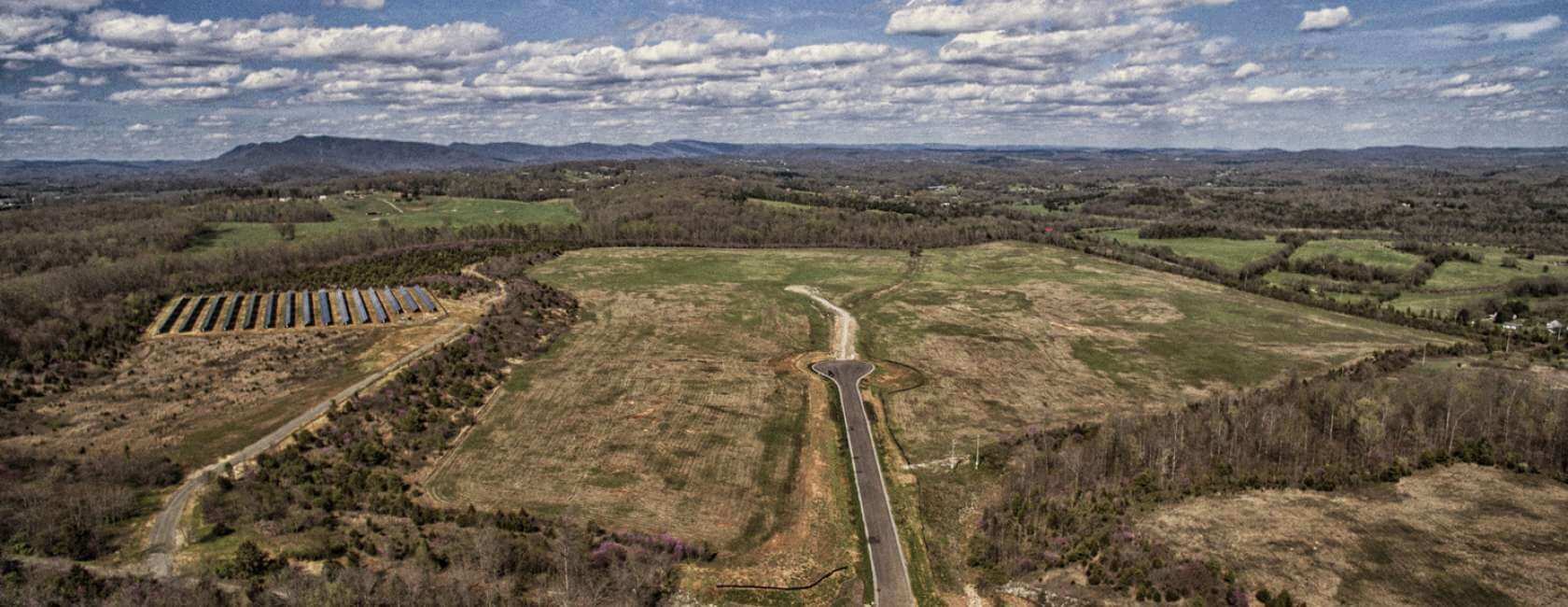 Aerial view of a wide, open grassy field with a road leading into it, surrounded by wooded areas and rolling hills under a partly cloudy sky. A set of solar panels is visible on the left side.