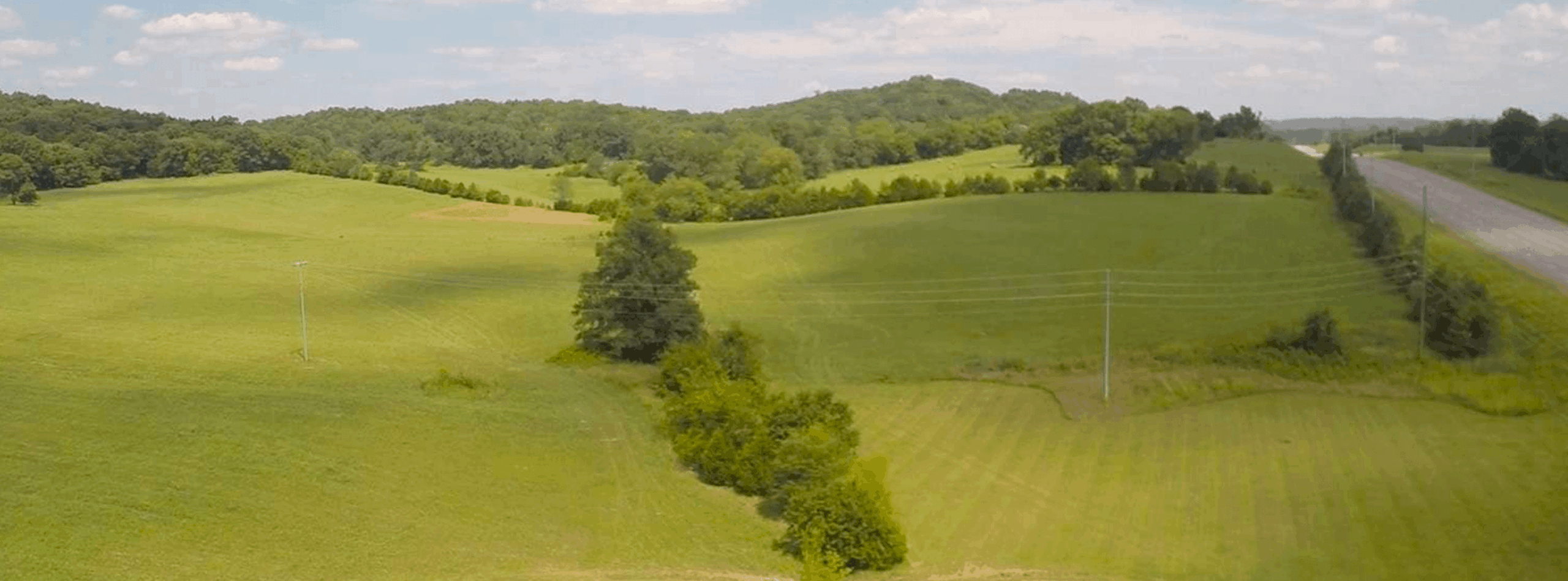 Rolling green hills under a partly cloudy sky, with scattered trees, power lines, and a road running along the right side of the landscape.