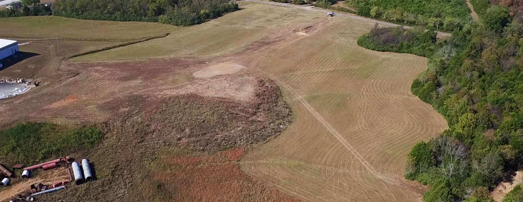 Aerial view of a large, grassy, recently mowed open field bordered by trees, with some scattered industrial pipes and materials in the lower left corner and a road running along the upper edge.