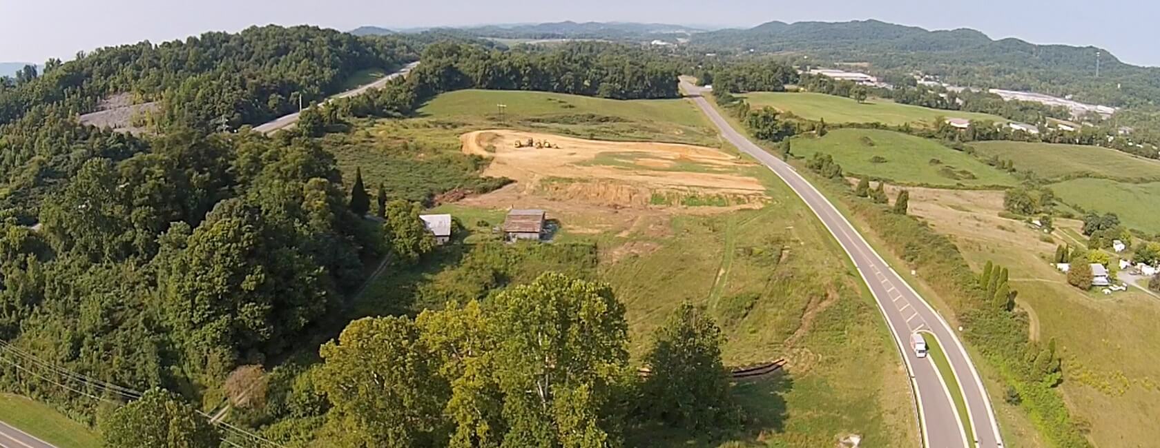 Aerial view of a rural landscape with a winding road, green fields, scattered trees, a few small buildings, and hills in the background under a clear sky.