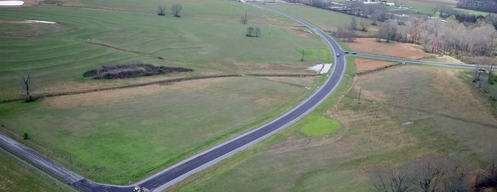 Aerial view of a rural landscape with green fields, a few trees, and a winding road intersecting through the area under a cloudy sky.