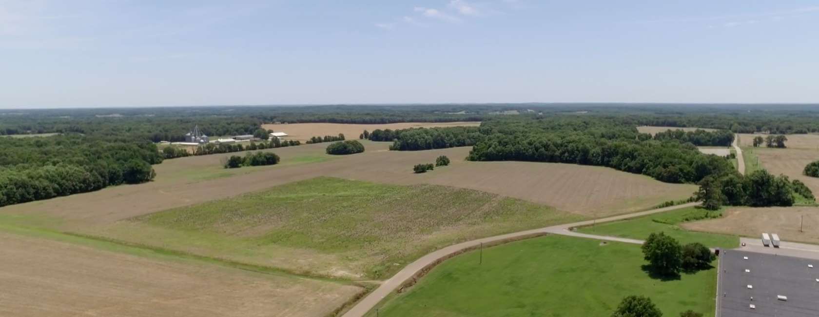 Aerial view of a rural landscape with open fields, patches of trees, a winding road, and a building in the lower right corner under a clear blue sky.