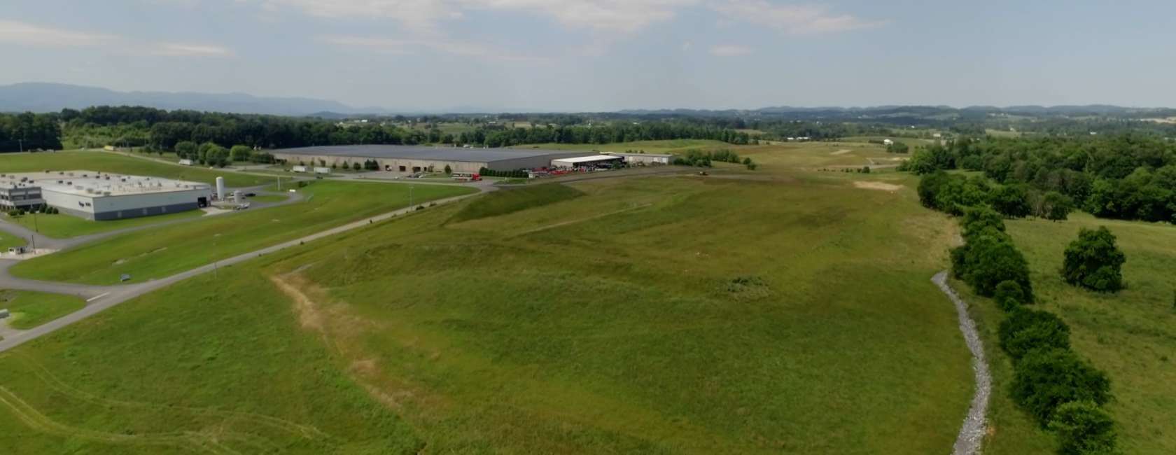 Aerial view of a large grassy field with scattered trees, industrial buildings, roads, and distant hills under a partly cloudy sky.