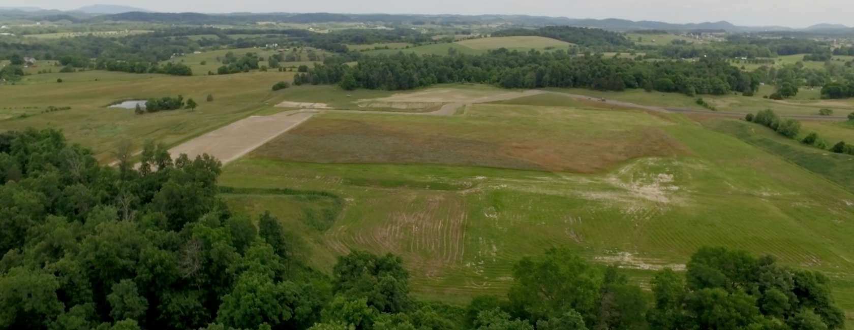 Aerial view of a large, open field with patches of grass and dirt, surrounded by trees and rolling hills under a cloudy sky in a rural landscape.