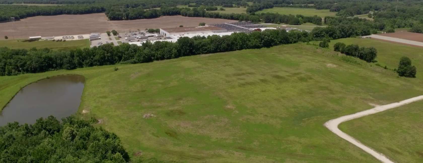 Aerial view of a large grassy field with a pond on the left, a white gravel road on the right, and a forested area separating the field from an industrial complex and parking lot in the background.