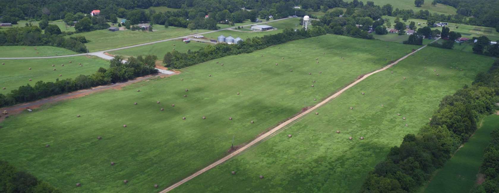 Aerial view of a large, green rural field dotted with hay bales, a dirt road running through it, and trees, houses, and farm buildings in the background.
