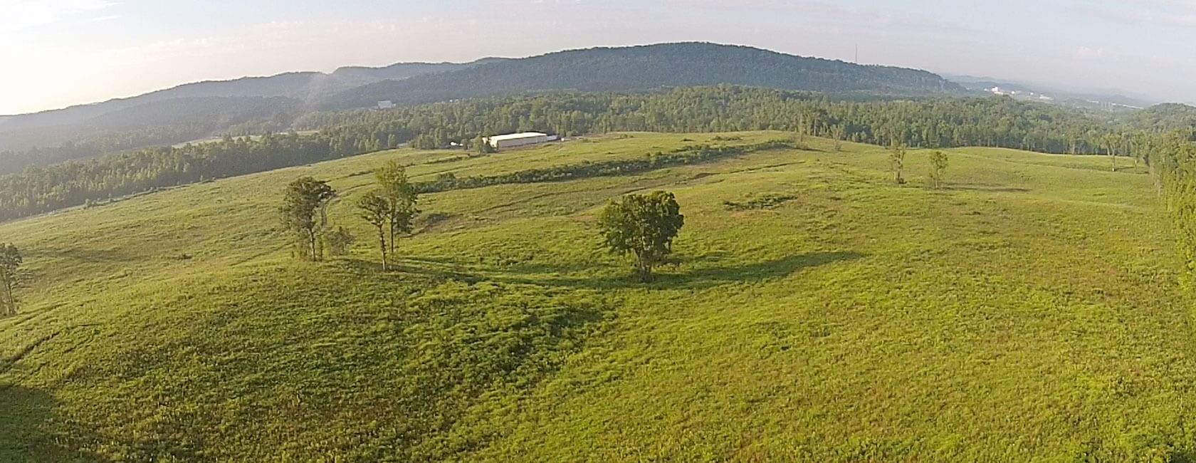 Rolling green hills with scattered trees and a few buildings in the distance, set against a backdrop of forested mountains under a partly cloudy sky.