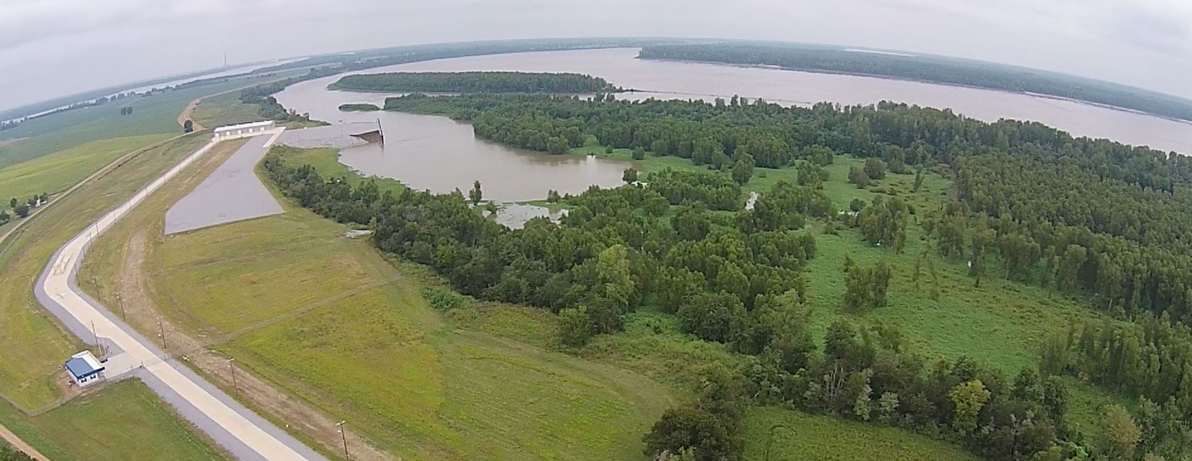 Aerial view of a dam structure next to a river, surrounded by green fields, dense trees, and a winding road; the river curves through the landscape under an overcast sky.