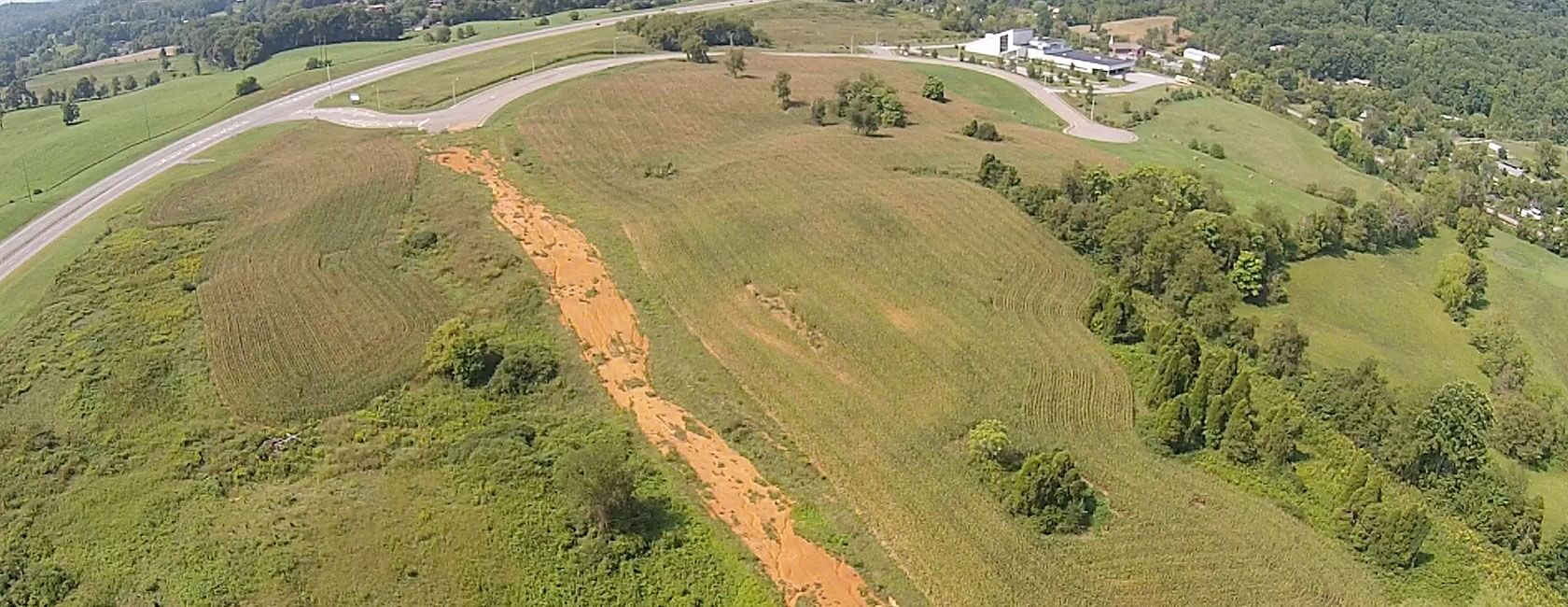 Aerial view of a grassy, hilly landscape with a large, exposed strip of reddish-brown soil running diagonally through the center, surrounded by green fields, trees, and a road in the background.
