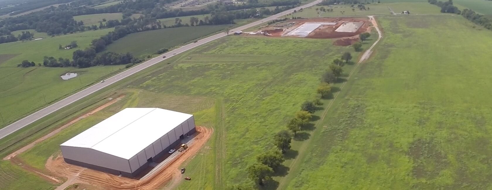 Aerial view of a large white warehouse beside a road, surrounded by green fields; construction activity and cleared land are visible further along the road.
