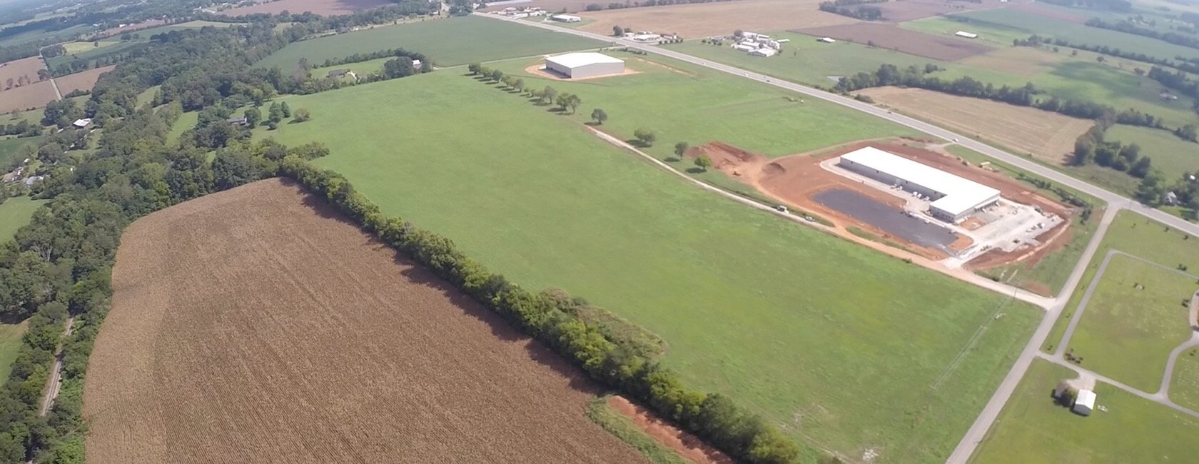Aerial view of a rural landscape featuring green fields, some farmland, a few scattered buildings, and roads intersecting the area under a clear sky.