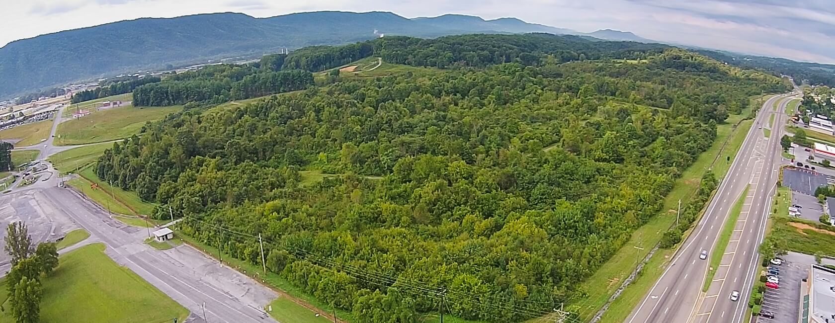 Aerial view of a dense green forest bordered by roads, parking lots, and distant mountains under a cloudy sky, with vehicles visible on the road to the right.