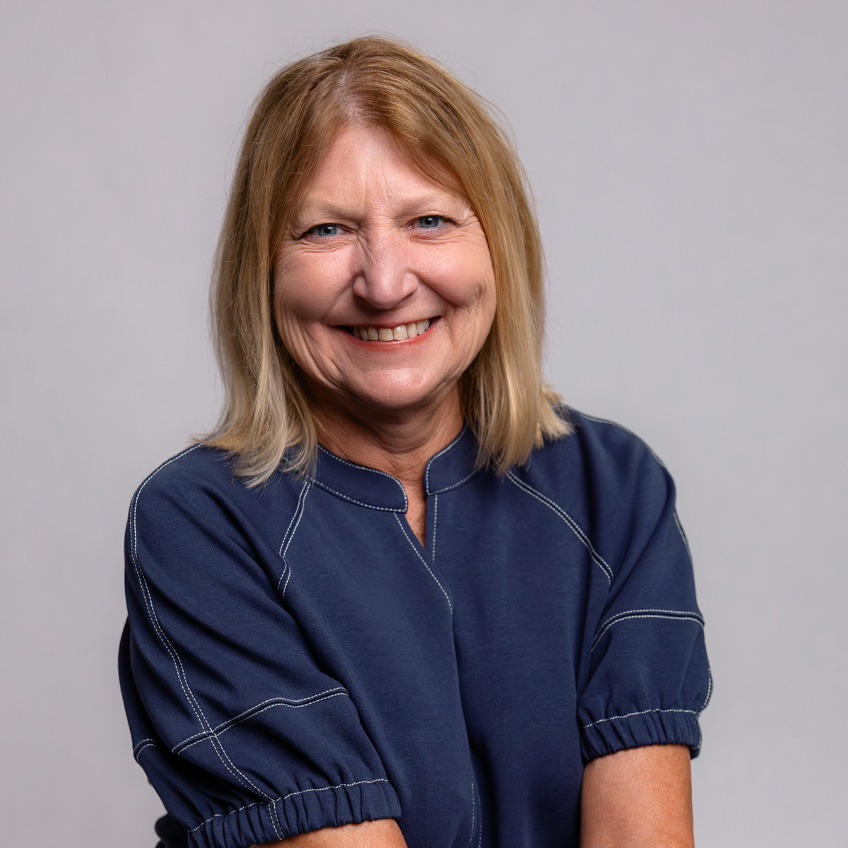 A smiling older woman with shoulder-length blonde hair wearing a navy blue top, standing against a plain light gray background.