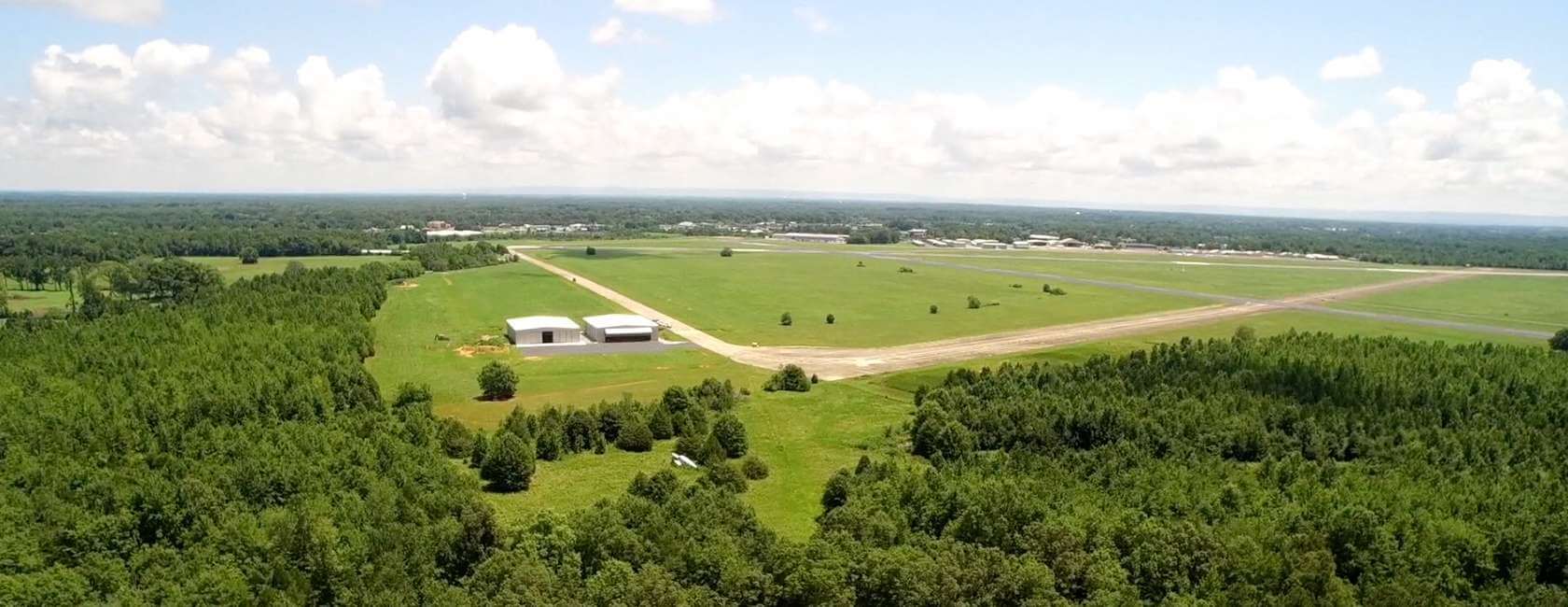 Aerial view of a rural airport surrounded by dense green forest, with runways, grassy fields, and a few small buildings under a partly cloudy sky.