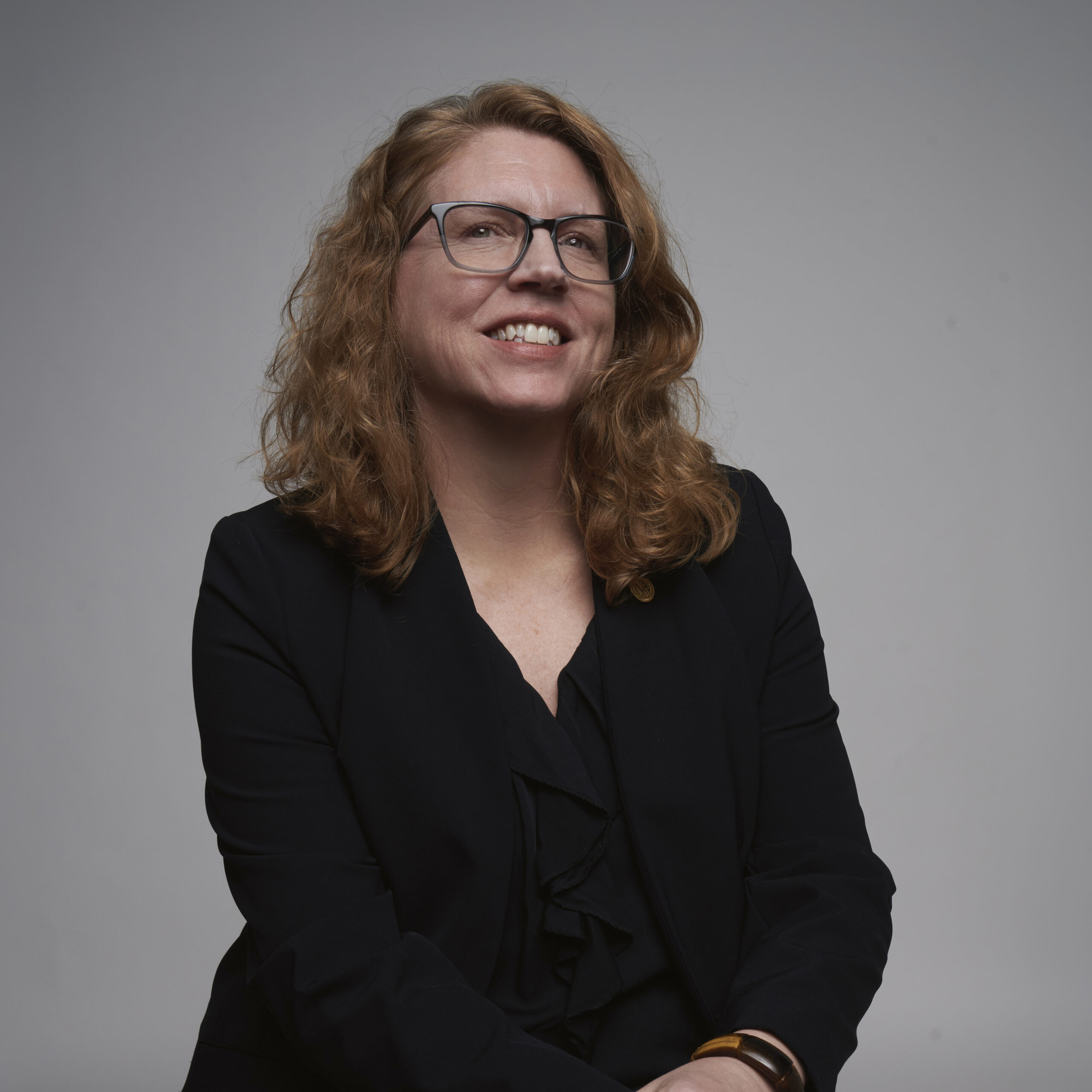 A woman with wavy, shoulder-length hair and glasses smiles while wearing a black blazer and shirt, seated against a plain gray background.