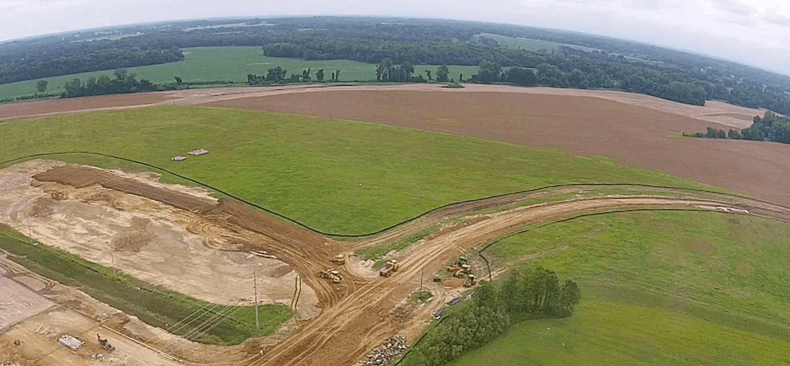Aerial view of a construction site in a grassy field, with dirt roads and earth-moving equipment. Surrounding areas include green fields, trees, and a wooded horizon under a cloudy sky.