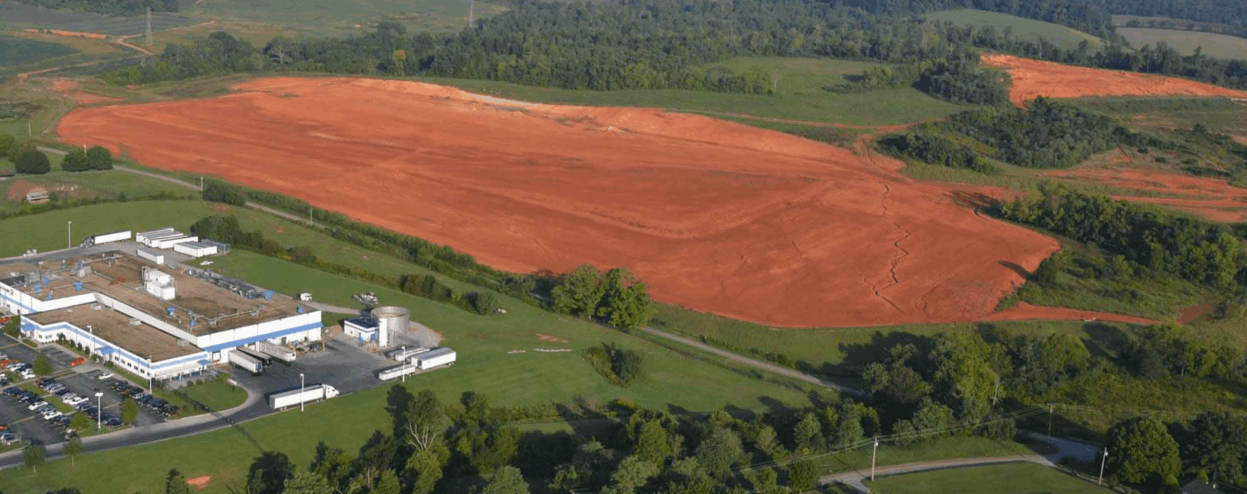 A large, cleared area of reddish soil sits next to green fields and a white industrial building, surrounded by trees and rolling hills under a clear sky.