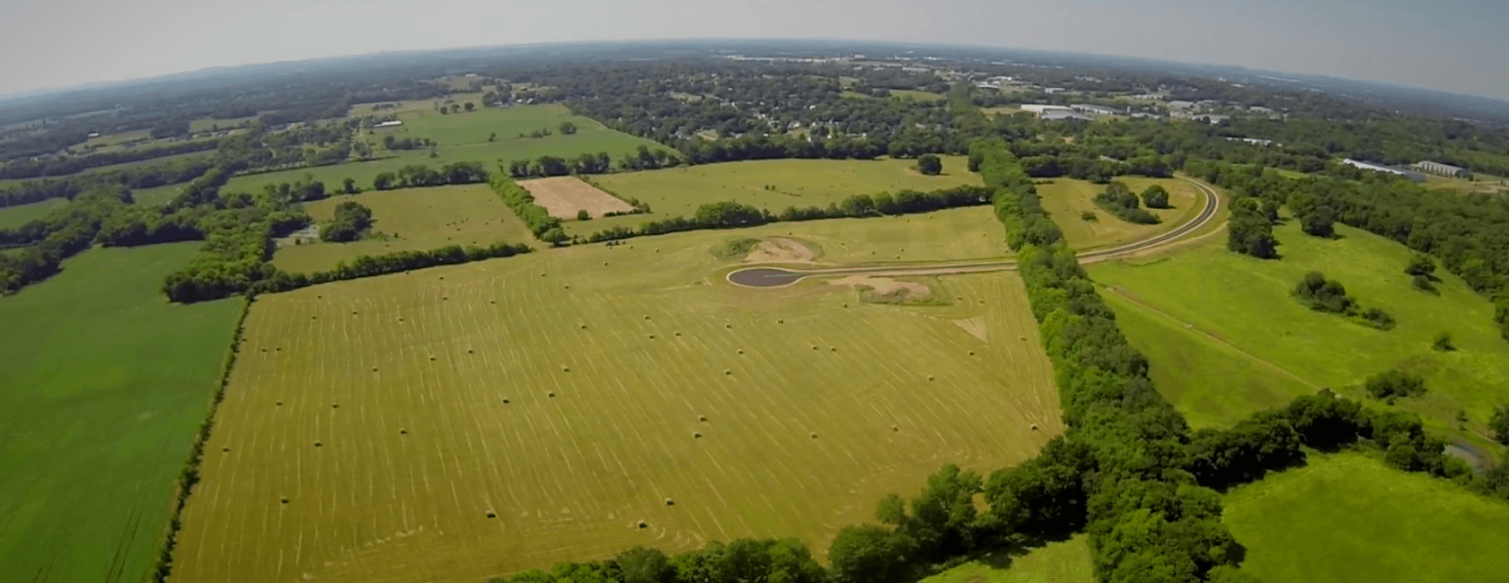 Aerial view of a large, green rural landscape with neatly cut fields, scattered hay bales, a winding road, and clusters of trees under a clear sky.