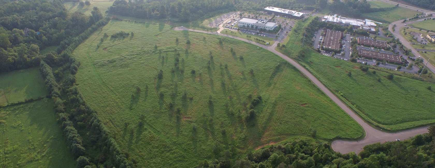 Aerial view of a large grassy field with scattered trees, bordered by a road and dense forest. Buildings with parking lots are visible at the top right, and the landscape appears lush and green.