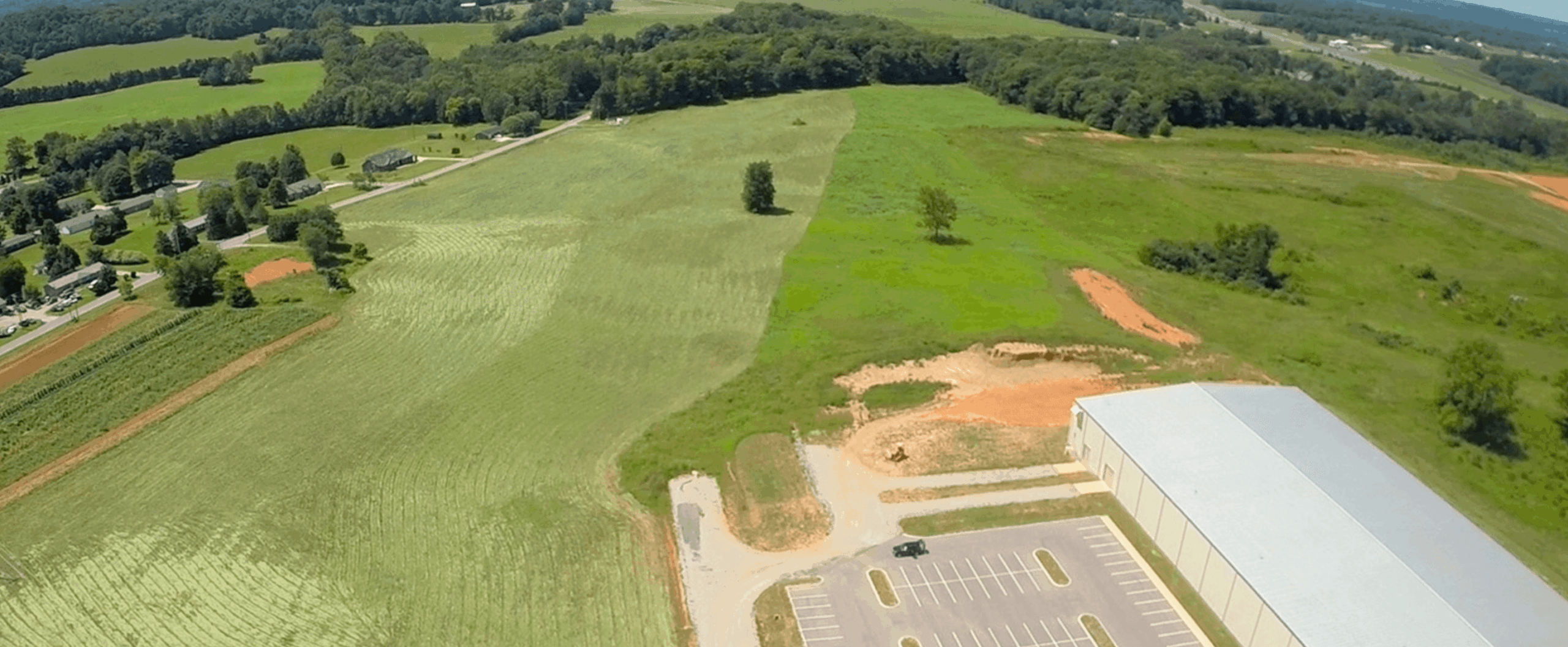 Aerial view of a rural landscape showing open green fields, a few scattered trees, a large beige building with a parking lot, and a road with houses along the edge of the image. Forested hills are visible in the distance.