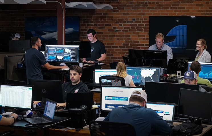 An office with several people working at computer desks. Multiple monitors display data and graphs. The room has exposed brick walls and aviation-themed decor. Some people are standing while others are seated, focused on their tasks.