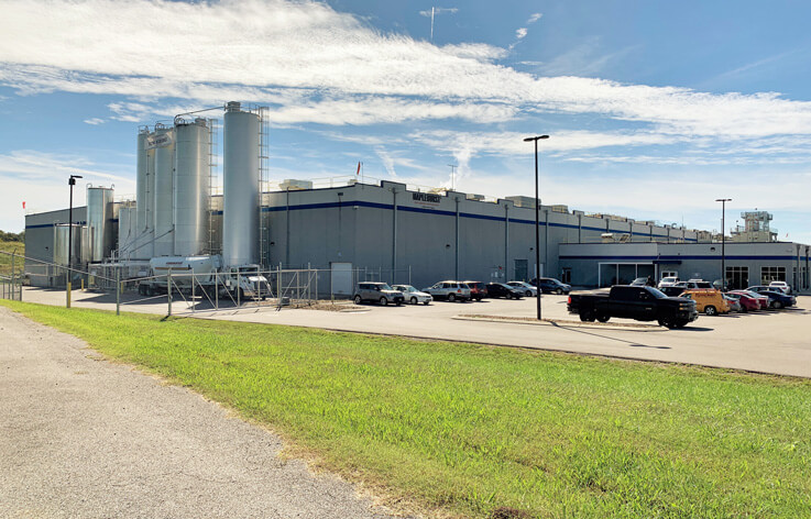 An industrial facility with multiple storage silos and a gray building sits under a blue sky. Several cars and trucks are parked in the lot, and green grass lines the road in the foreground.
