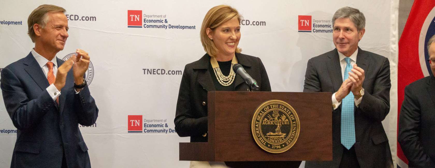 A woman speaks at a podium with the Tennessee state seal, while two men in suits stand on either side applauding. A backdrop features the TN Department of Economic & Community Development logos.
