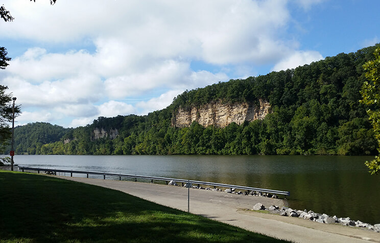 A calm river flows beside a tree-covered bluff with exposed rock ledges under a partly cloudy sky. A paved path and metal railing run alongside the river in the foreground.
