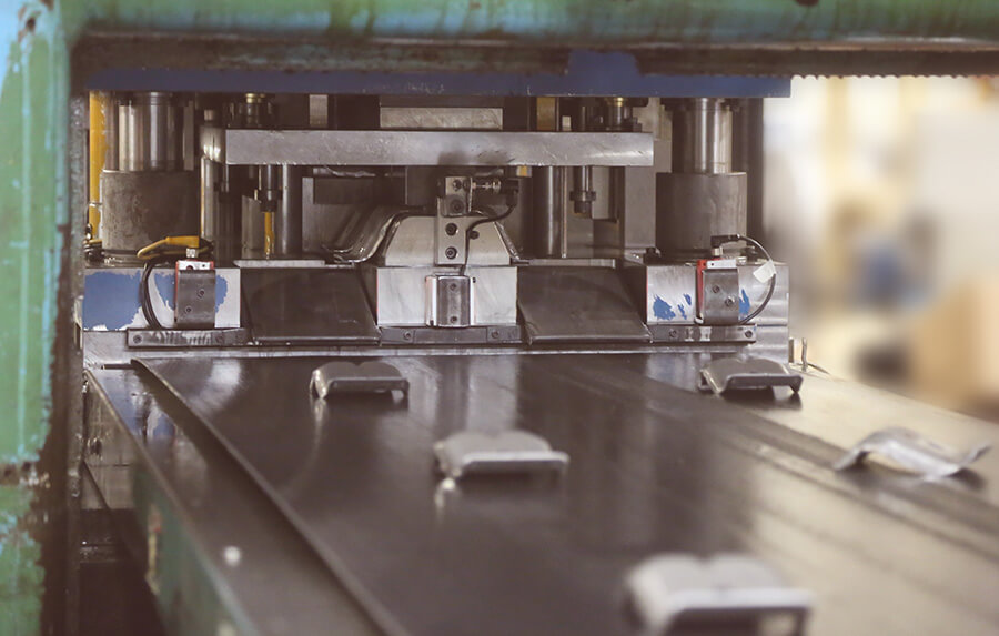 Close-up of an industrial machine stamping or shaping metal sheets, with several small metal parts visible on the conveyor belt. The setting appears to be a factory or manufacturing facility.