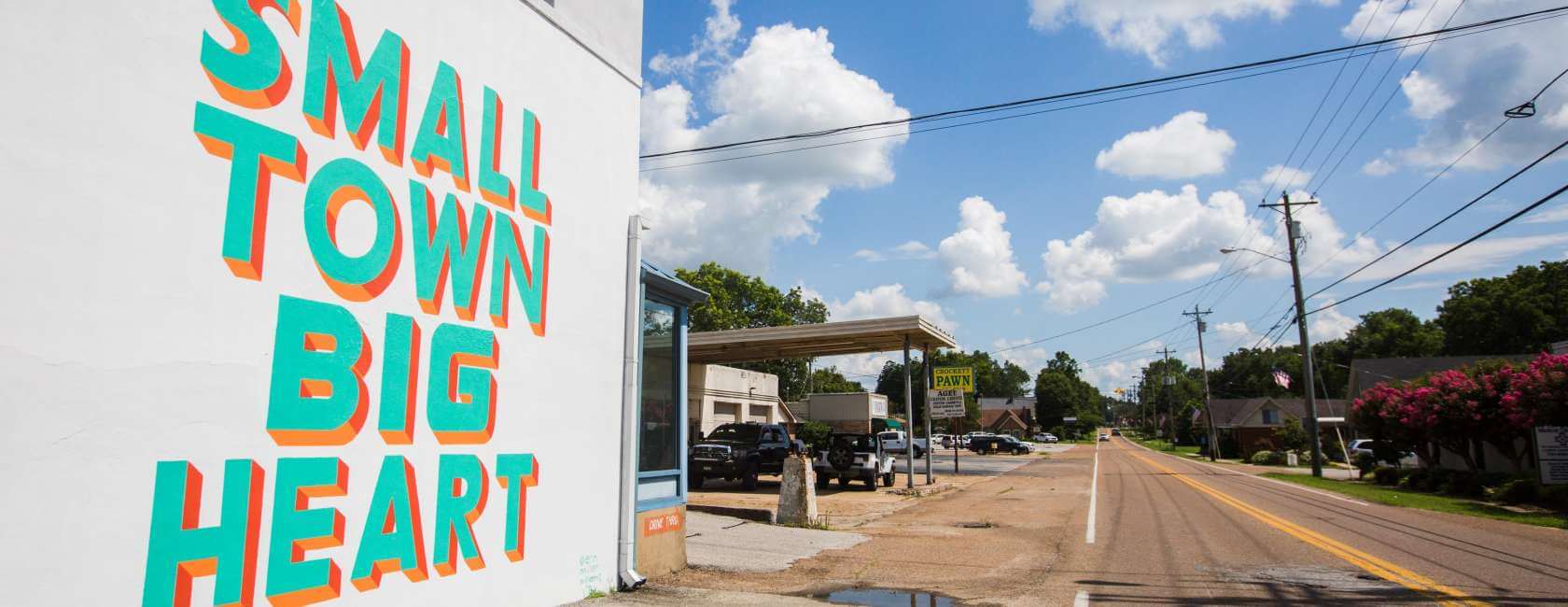 A colorful mural on a white wall reads “SMALL TOWN BIG HEART” beside a quiet main street lined with trees, parked cars, and a blue sky with fluffy clouds overhead.