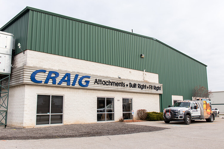 A large industrial building with a green metal roof and siding, labeled "CRAIG Attachments - Built Right, Fit Right." A branded pickup truck is parked outside near the entrance.