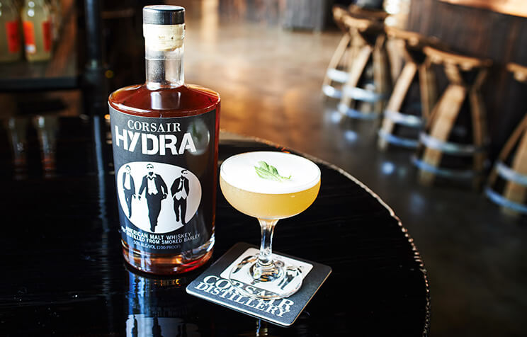 A bottle of Corsair HYDRA whiskey sits on a glossy black table beside a cocktail garnished with a leaf, with a Corsair-branded coaster underneath. Wooden bar stools and shelves are visible in the blurred background.