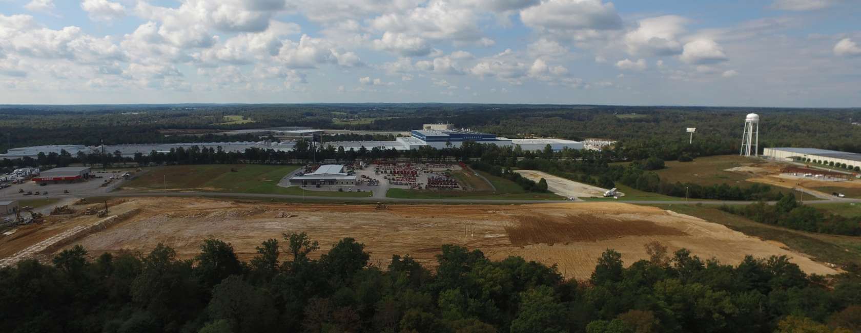 Aerial view of a construction site with cleared land in the foreground, buildings and parking lots in the middle, surrounded by trees, with a water tower and rolling landscape under a partly cloudy sky.