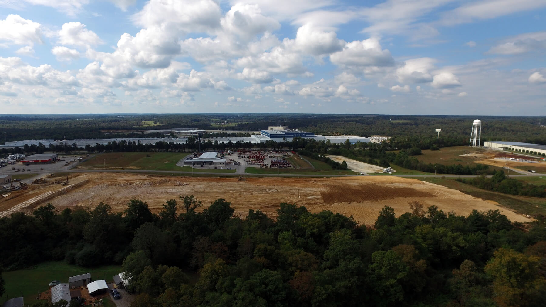 Aerial view of a semi-rural area with cleared land in the foreground, trees along the bottom, scattered buildings, a parking lot, and a large water tower under a sky with scattered clouds.