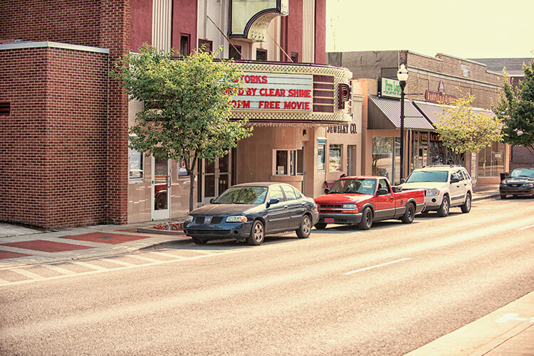 Three vehicles—a blue car, a red truck, and a white SUV—are parked along a small town street in front of businesses, including a theater with a marquee offering a free movie. Trees line the sidewalk.