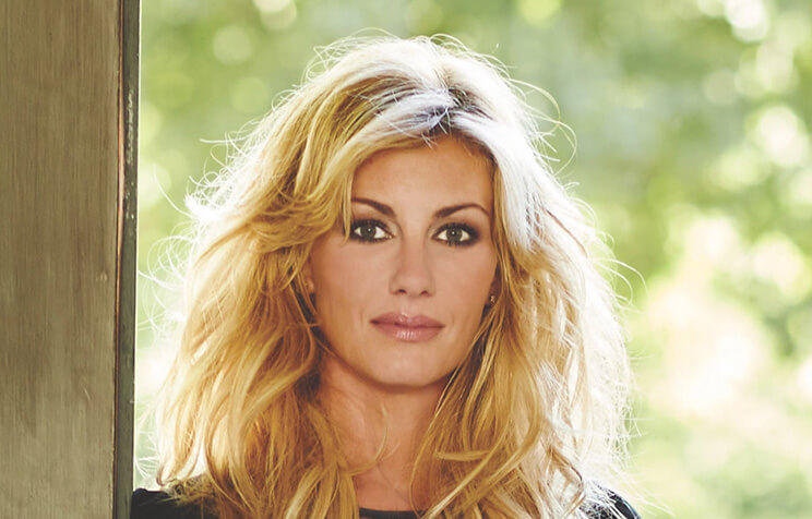 A woman with long, wavy blonde hair and dark eye makeup gazes directly at the camera, standing outdoors with green foliage blurred in the background.