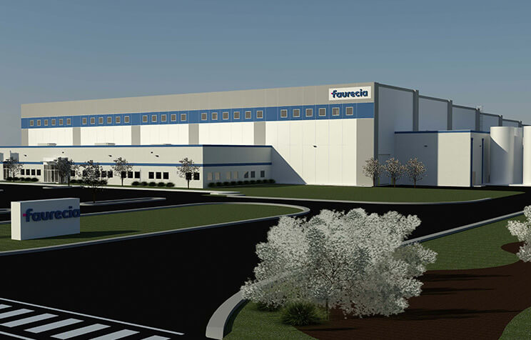 A large, modern industrial building with the Faurecia logo, surrounded by a paved road, parking area, grass, and a few small trees under a clear sky.