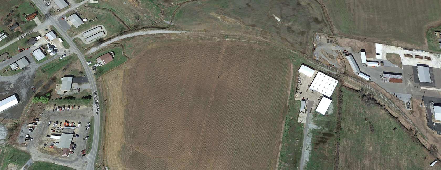Aerial view of a rural area showing a large, empty brown field surrounded by roads, several buildings, parking lots, and patches of grass and trees. Industrial and residential structures are visible around the field.