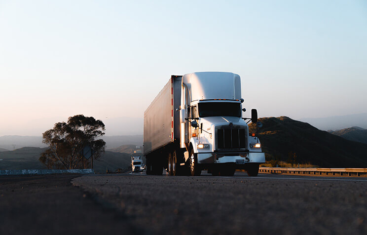 A white semi-truck drives along a highway at dusk with mountains and trees in the background. Another truck is visible further down the road. The sky is clear and softly lit.