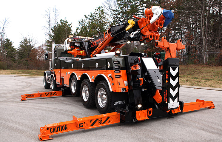 A large, heavy-duty tow truck with bright orange stabilizer arms extended on both sides and rear, parked on pavement; trees and overcast sky are in the background.