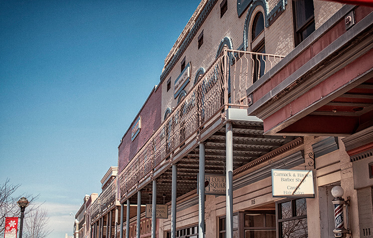 A row of historic brick buildings with metal balconies and business signs, under a clear blue sky. The architecture features arched windows and decorative trim, evoking a vintage, Old West feel.