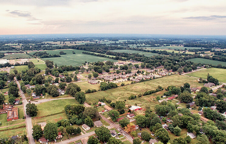 Aerial view of a rural town with scattered houses, tree-lined streets, open fields, and farmland stretching out to the horizon under a partly cloudy sky.
