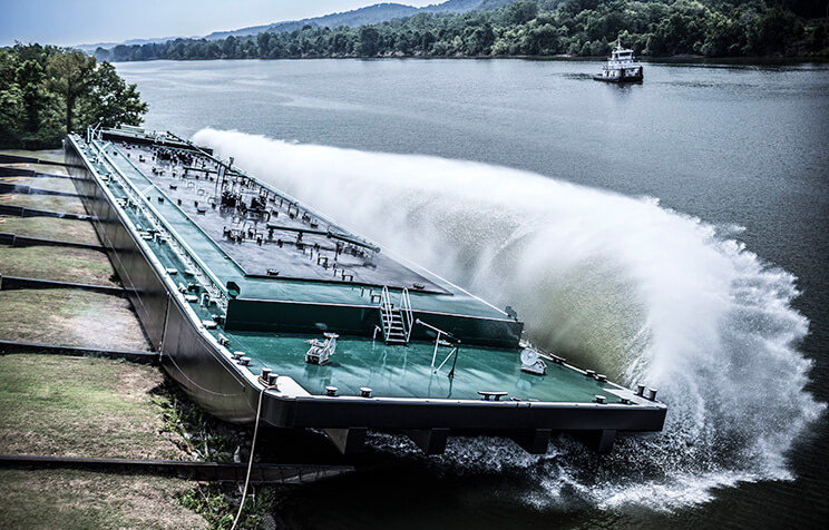 A large barge slides sideways into a river, creating a massive splash and waves, while another vessel is seen in the distance; trees line both banks under a cloudy sky.