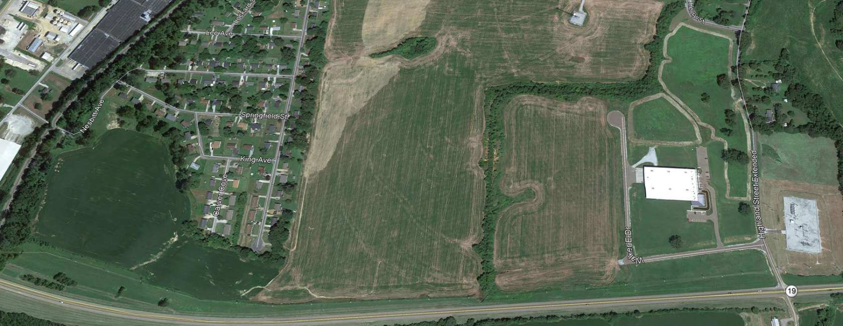 Aerial view of a suburban neighborhood with houses and streets on the left, open farmland in the center, and the Walker Industrial Site with a large building and parking lot on the right, bordered by roads and green areas.