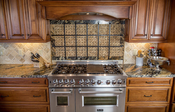A stainless steel gas range with dual ovens is set in a kitchen with wooden cabinets, granite countertops, and a decorative tile backsplash. Kitchen utensils, a knife block, and a colander sit on the counters.