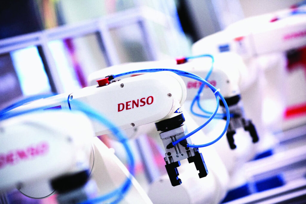 Close-up of white robotic arms with blue cables and the word "DENSO" printed on them, operating in a modern industrial or manufacturing environment.